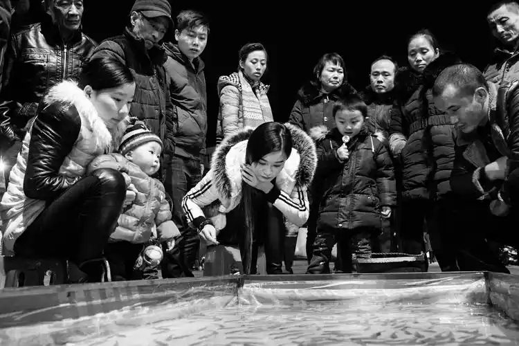 一群人正围在一起,看一个美女玩钓鱼游戏,美女专注的表情和围观的人