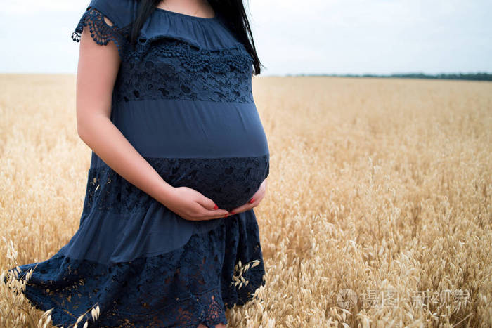 照片 年轻漂亮的9个月孕妇穿着蓝色连衣裙,抱着肚子,在夏日户外的燕麦