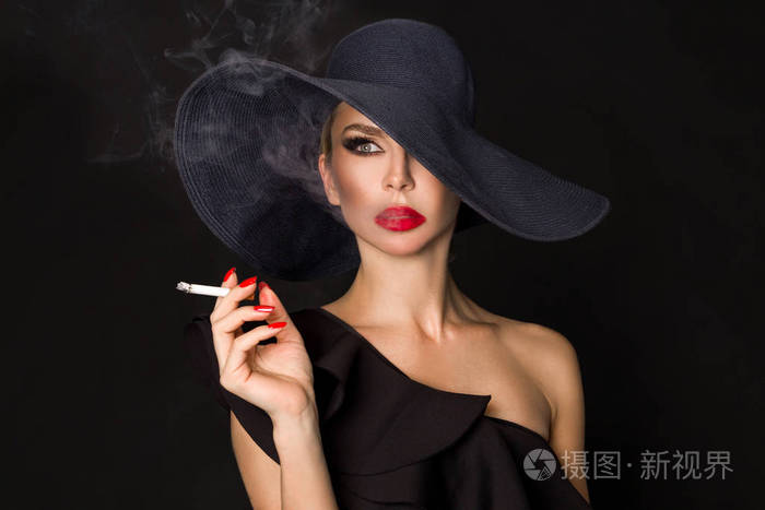 优雅的女人,黑党徽与香烟在手拿着帽子.在黑色背景上